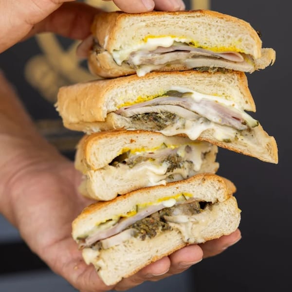 A hand holds a stack of four toasted Cuban sandwiches with meat, cheese, mustard, and pickles.
