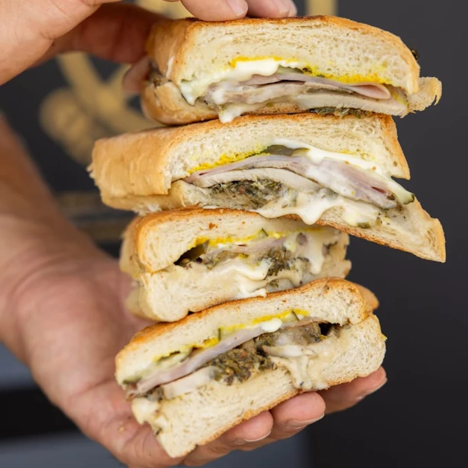 A hand holds a stack of four toasted Cuban sandwiches with meat, cheese, mustard, and pickles.