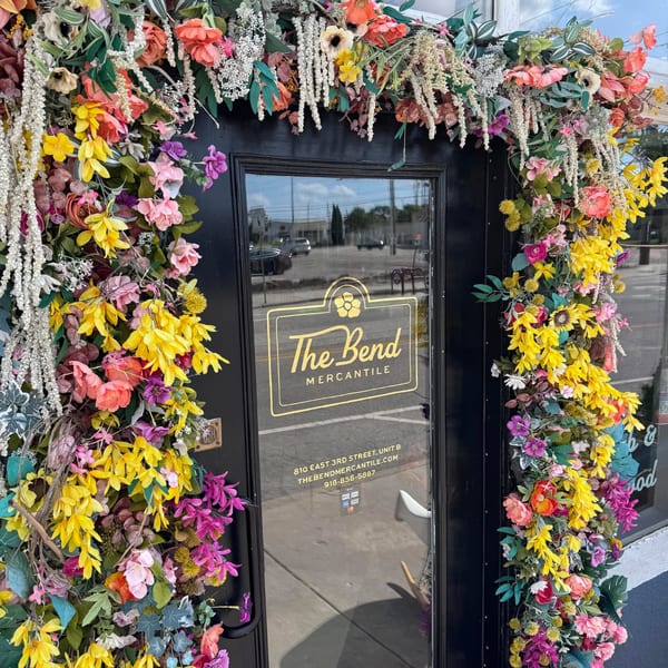 The Bend Mercantile storefront, adorned with vibrant artificial flowers around a black door and window.