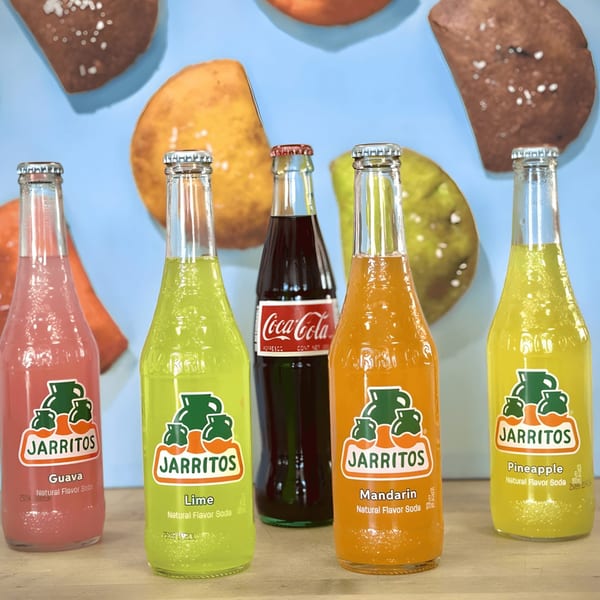 Five colorful Jarritos and one Coca-Cola bottle stand against a blue background with empanada art.