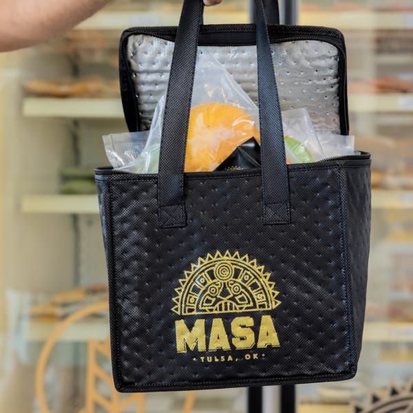 Hand holding a black MASA branded insulated bag filled with packaged empanadas and sauces.