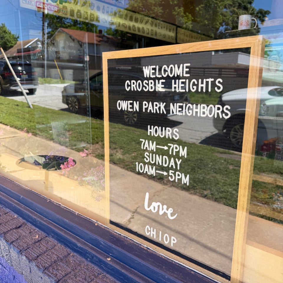 A window sign displays "Welcome Crosbie Heights Owen Park Neighbors" and store hours, reflecting in the glass.