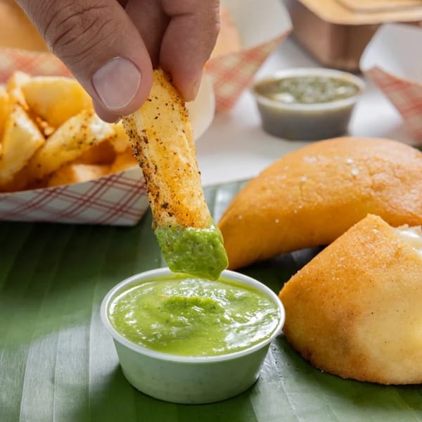Hand dipping a seasoned potato wedge into green sauce, with MASA Empanadas in the background.