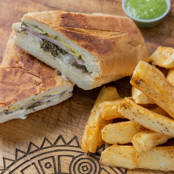 Cuban sandwich with melted cheese and green sauce, served with seasoned yucca fries on a wooden board.
