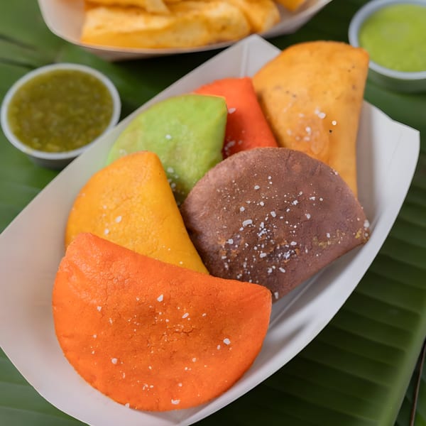 A white tray holds colorful MASA Empanadas in orange, green, yellow, and brown, with green sauce.